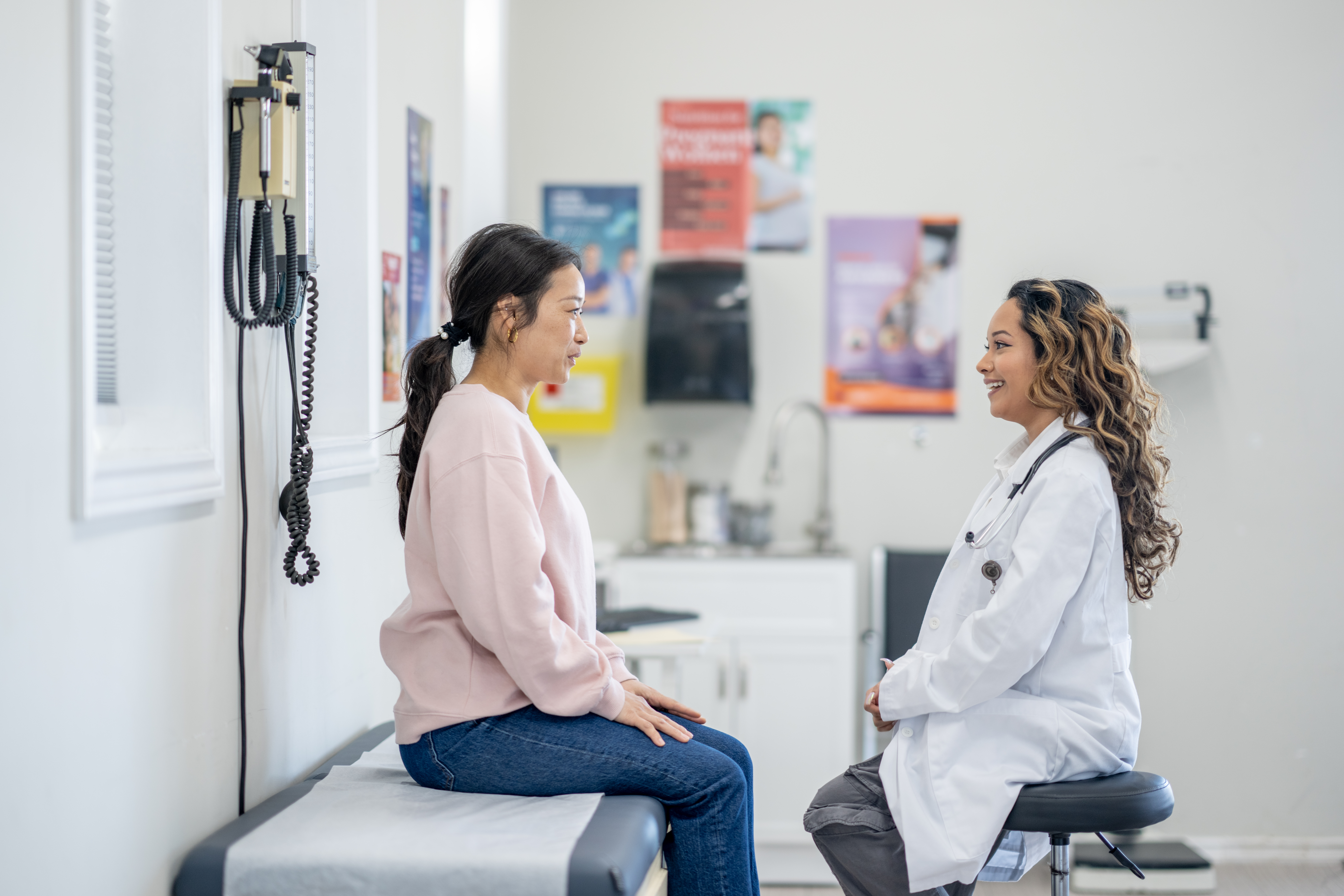 A woman sits up on an exam table as she talks with her female doctor about her health concerns.