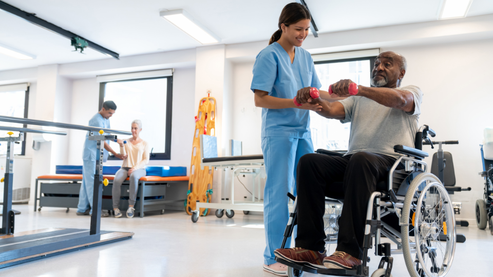 Male patient in a wheelchair working out his arms using dumbbells with the help of a physical therapist