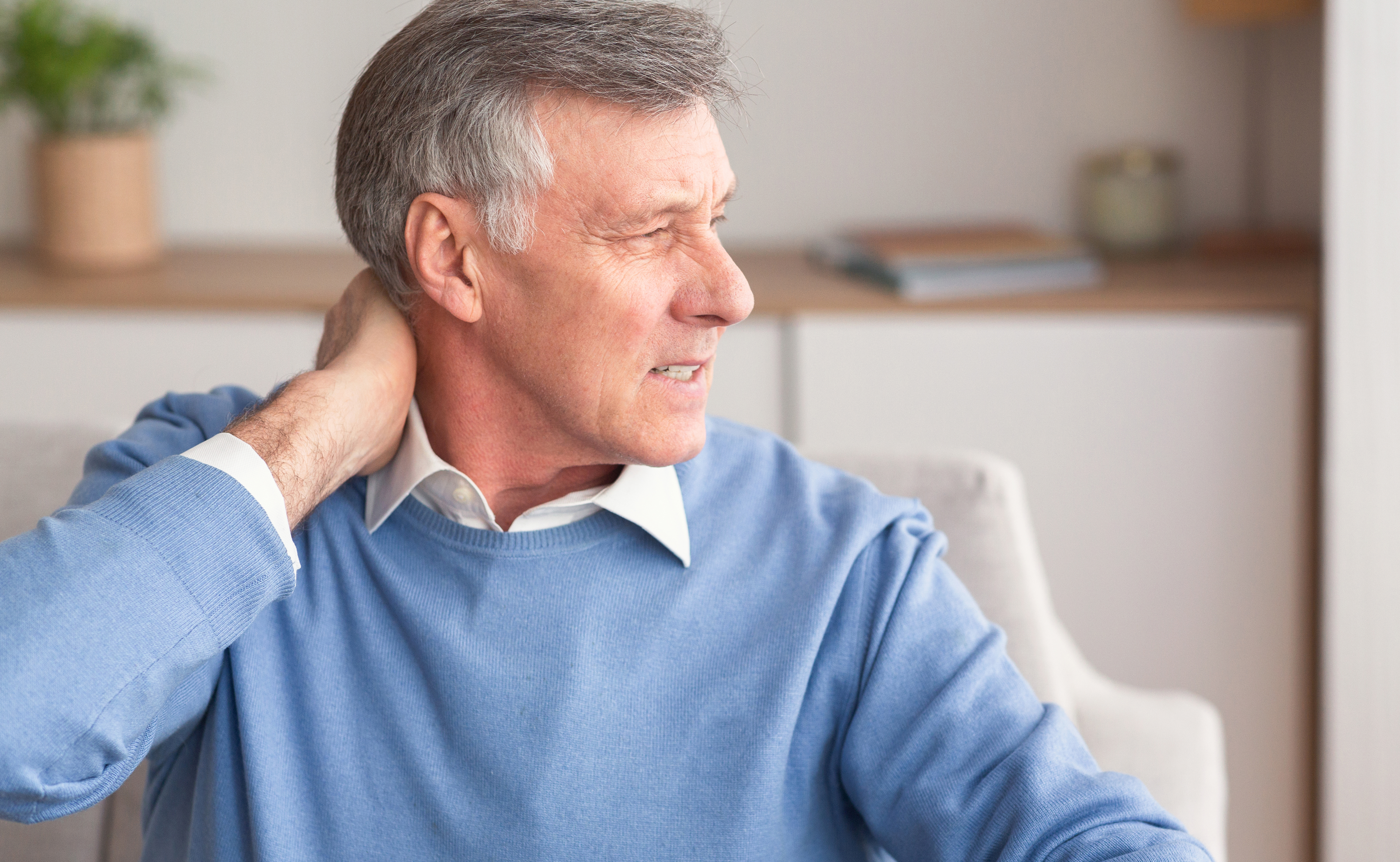 An older man in a blue sweater grabs at his neck in pain