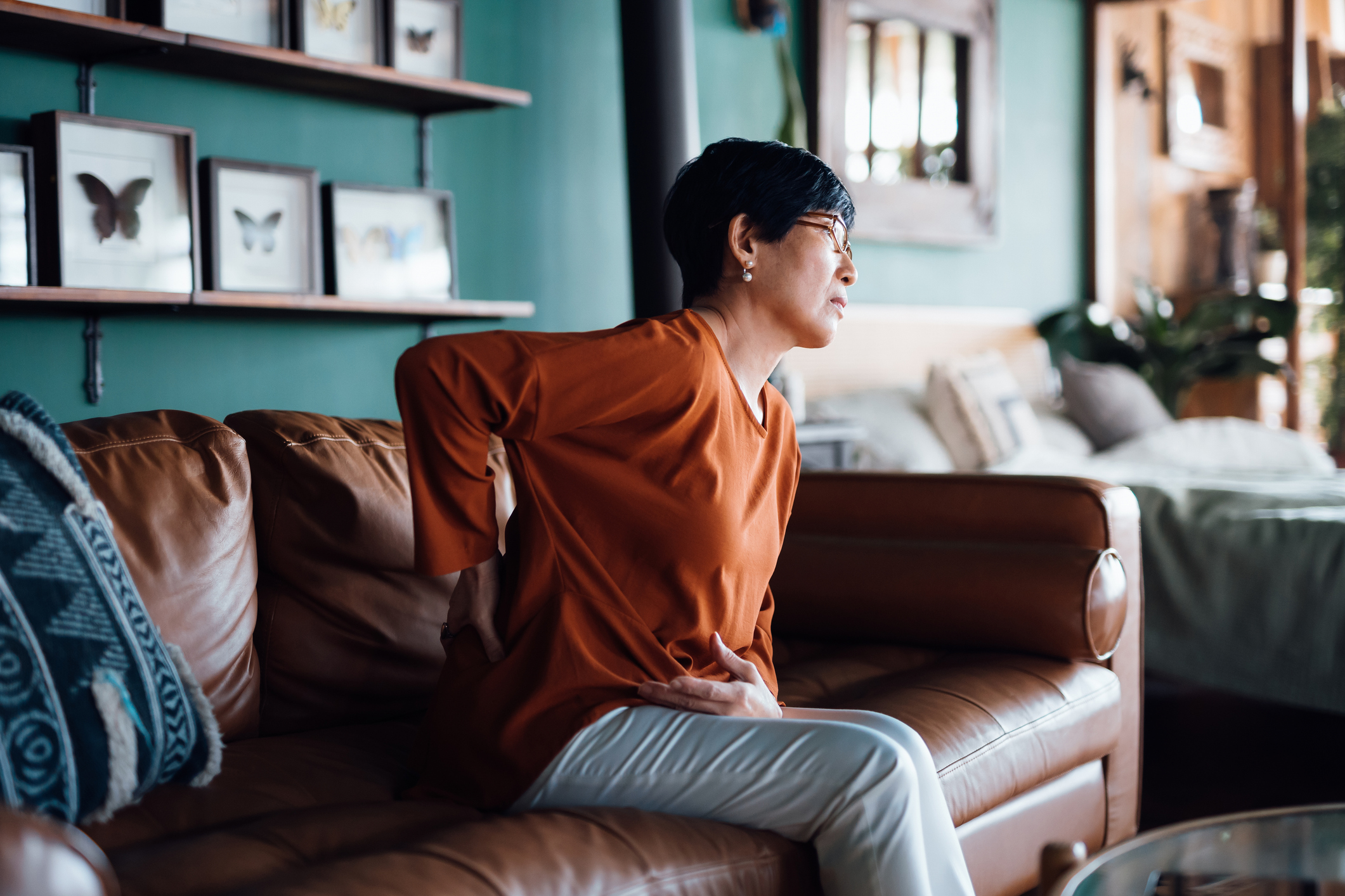 an older woman sits on a couch and grabs her lower back in pain