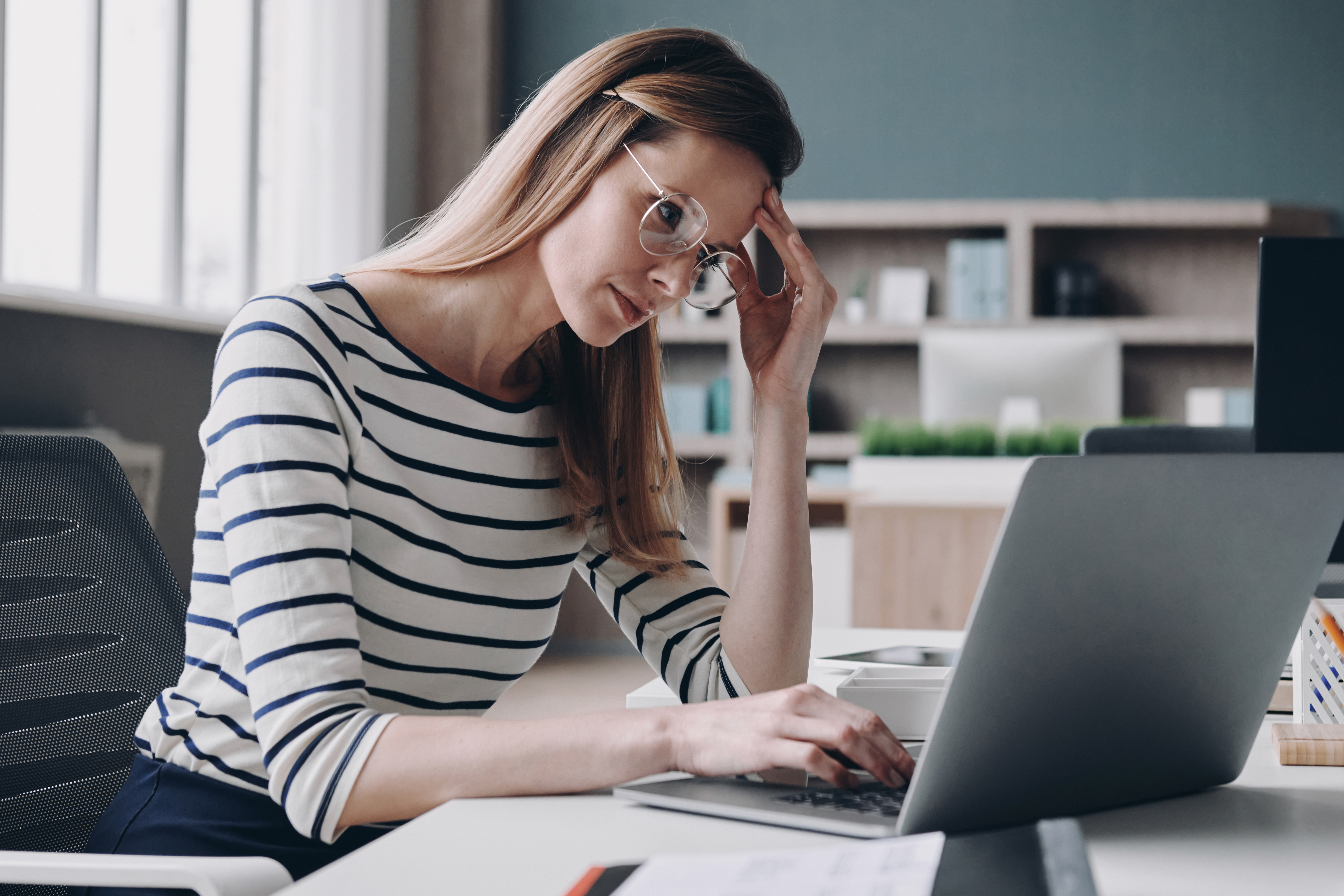 Tired young businesswoman using laptop and holding head in hand while working in the office