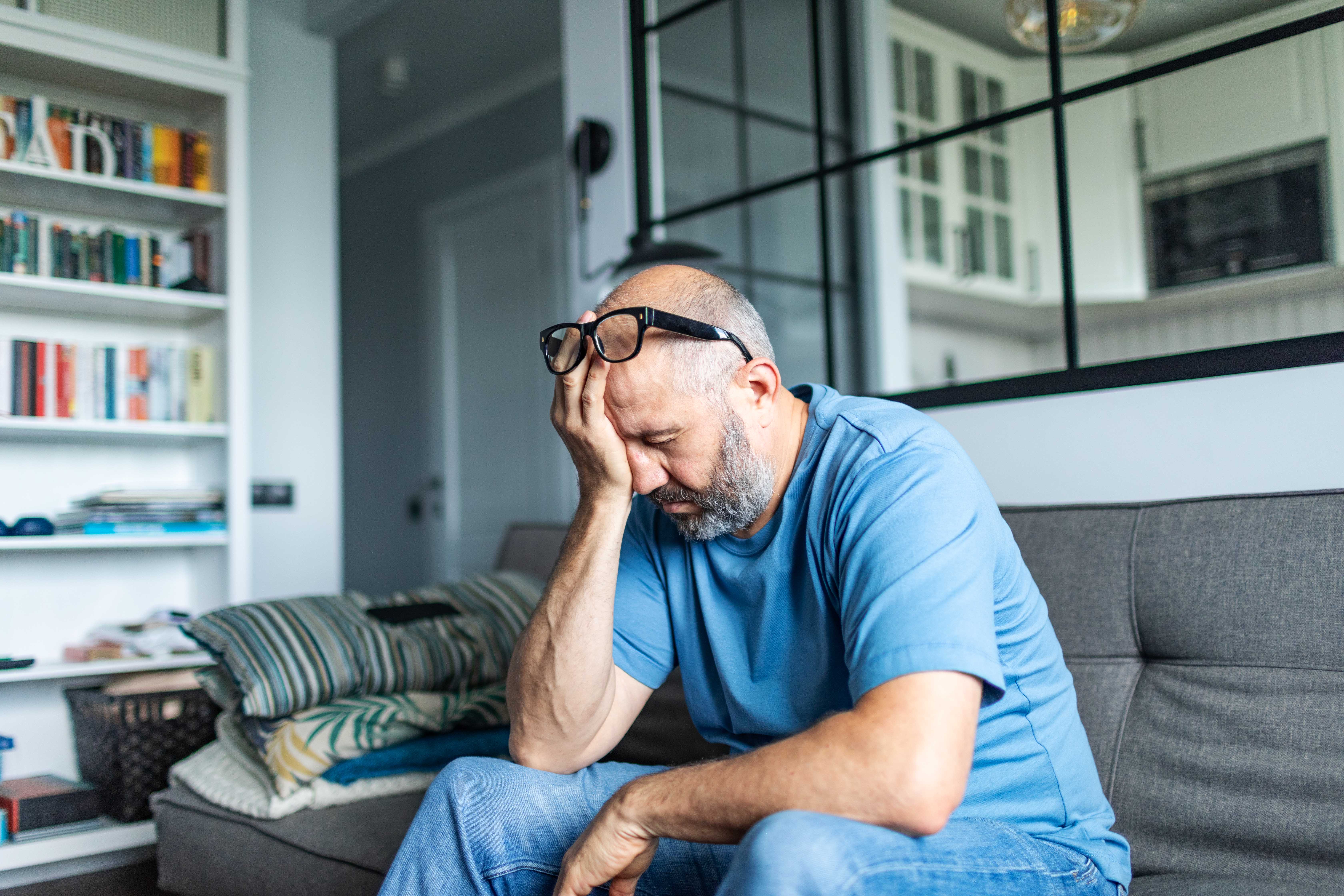 a middle aged man sits on a sofa with his head in his hand, showing distress
