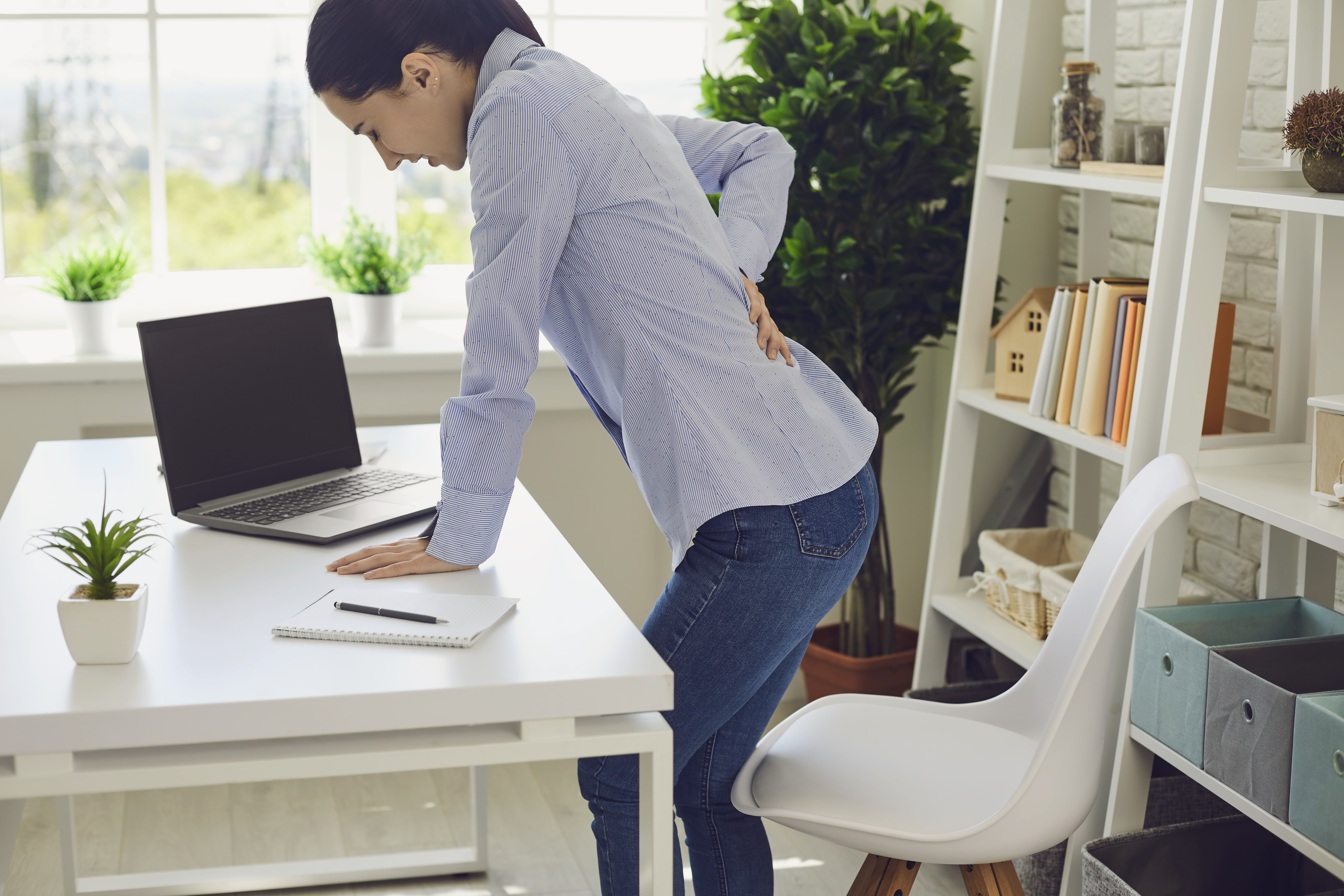 A woman stands up at her desk and holds her lower back in pain